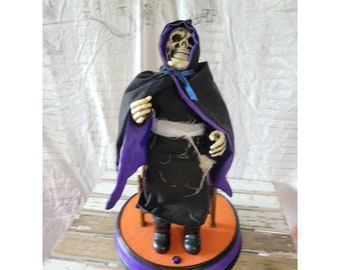 May include: Animated Halloween decoration of a skeleton wearing a black robe with a purple lining and a blue ribbon. The skeleton is sitting in a chair and has a white bandage around its waist. The decoration is on a round base with an orange center and a purple rim.