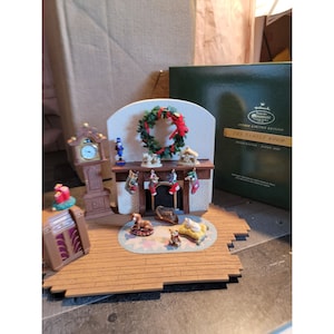 May include: A hand-crafted Christmas village scene featuring a fireplace with stockings, a clock, a nutcracker, a wreath, and a nativity scene. The scene is set on a wooden floor and includes a small dog and cat figurine. The box reads "The Family Room, Studio Limited Edition, Handcrafted, Dated 2002."