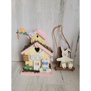May include: A decorative wooden Easter-themed set featuring a pastel pink and yellow birdhouse with the text "Bunny Inn" and a bunny on a swing. The birdhouse has a carrot and bird accents.