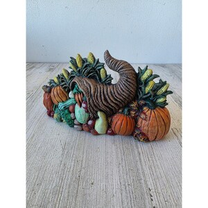 May include: A decorative cornucopia overflowing with autumn harvest items. The horn is brown and textured, surrounded by pumpkins, gourds, corn, and other vegetables in shades of orange, green, and yellow. A festive centerpiece.