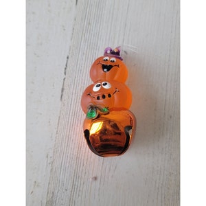 May include: A stack of three orange pumpkins with painted faces, the top pumpkin wearing a purple hat. The bottom pumpkin is a bell with a green leaf.