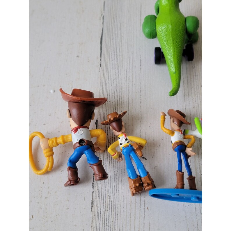 May include: A collection of Toy Story character figurines, including Woody, Buzz Lightyear, and Rex the dinosaur. Woody figures are in cowboy attire with brown hats, yellow shirts, blue pants, and brown boots. Rex is green with black wheels.