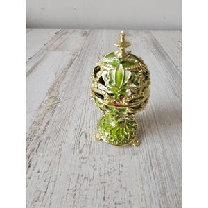May include: An ornate, decorative egg-shaped object with a gold-colored filigree design and green enamel accents. The egg features a detailed floral pattern and is adorned with small red jewels. The base and top are also gold, creating a luxurious appearance.