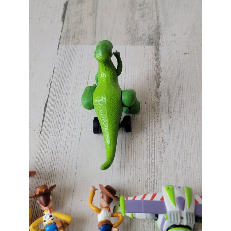 May include: A green toy dinosaur with black wheels, part of a collection of Toy Story character figurines. The dinosaur is positioned on a light-colored wooden surface, with other character figures visible.