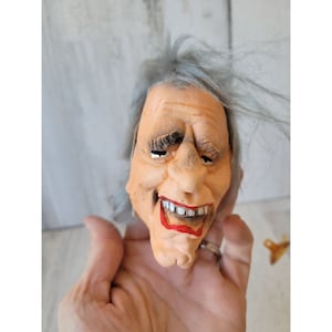 May include: A close-up of a rubber mask with a white wig. The mask has a realistic face with a wide smile and painted teeth. The mask is a light skin tone with a red mouth and gray hair.