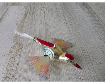 Può includere: Un ornamento a forma di uccello in vetro con corpo color argento metallizzato e testa e dorso rossi. L'uccello presenta ali traslucide a ventaglio e una coda in fibre sintetiche bianche, arricchita da dettagli di vernice maculata rossa, gialla e blu.