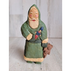 May include: A hand-carved wooden Santa Claus figurine with a green robe, white beard, and a red and blue toy soldier in his hand. He is holding a brown teddy bear in his other hand.