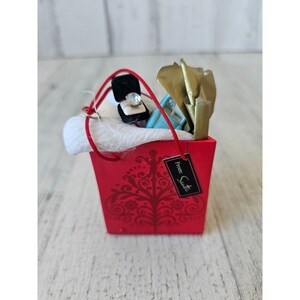 May include: A red shopping bag with a white bow and a gift box with a ring inside. The bag has a tag that says "From: Santa".