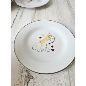 Könnte beinhalten: Ein weißer Keramikteller mit silbernem Rand, auf dem ein Cartoon-Rentier mit Pfeil und Bogen und das Wort "Cupid" in roten Buchstaben abgebildet ist. Um das Rentier herum sind kleine rote Herzen verstreut.
