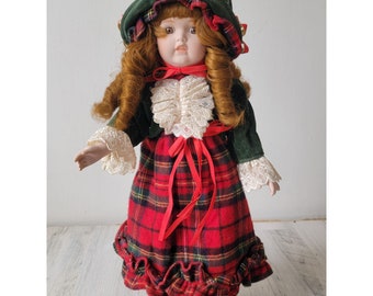 Op de afbeelding: Een porseleinen pop die een groen fluwelen jasje, een wit kant blouse en een rood-zwarte geruite rok met een rood lint draagt. De pop heeft rood haar, bruine ogen en een groene hoed met rode versiering en belletjes.