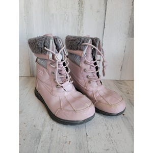 May include: A pair of pink winter boots with gray faux fur trim and laces. The boots have a black sole and the brand name "UGG" is visible on the side. The boots are designed for cold weather.