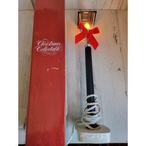 May include: A red box with the text "Christmas Collectable" on it. Next to the box is a black and silver lamp post with a red bow and a white cord. The lamp post is lit with a yellow light bulb.