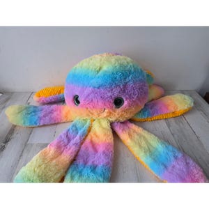 May include: A large, plush octopus toy in a rainbow of colors, including yellow, orange, pink, purple, and green. The stuffed animal has a smiling face with large, dark eyes and eight long, soft tentacles.