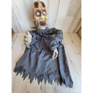 May include: A Halloween zombie decoration with a pale face, open mouth, and glowing white eyes. It wears a tattered grey robe with blood-like stains and a jagged hemline. The overall design creates a chilling and spooky atmosphere.