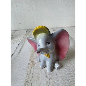 Könnte beinhalten: Eine Keramikfigur von Dumbo, dem Elefanten. Die Figur ist hauptsächlich grau mit rosa Ohren, einem gelben Hut und einer gelben Fliege. Der Elefant sitzt mit ausgebreiteten Ohren da. Ein klassischer Disney-Charakter.
