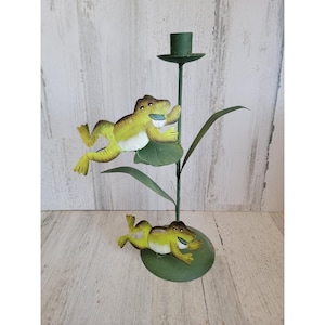 May include: A green metal candle holder featuring two yellow and green frog figurines. The frogs are positioned on lily pads and the candle holder has a single candle cup. The design is whimsical and decorative.