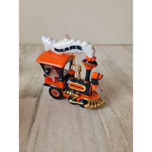 May include: A ceramic Christmas ornament shaped like a train with a Santa Claus figure in the cab. The train is orange, black, and gold with the words "Bears" and "2005" on it.