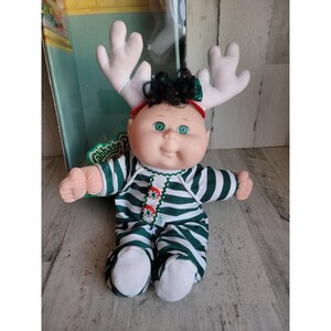 Può includere: Bambola Cabbage Patch Kids con tutina a righe verdi e bianche e fascia con corna di renna. La bambola ha gli occhi verdi e i capelli neri. La bambola è seduta davanti alla sua confezione originale.