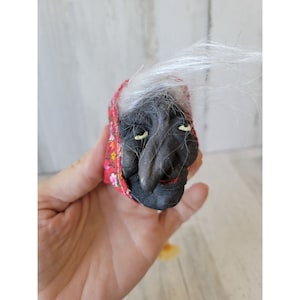 May include: A small, handmade doll head with a black face, white hair, and a red and white floral patterned fabric covering the head. The doll has a large nose and closed eyes.