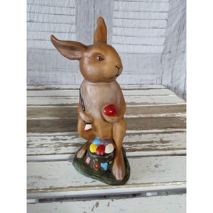May include: A brown ceramic Easter bunny figurine holding a paintbrush and a red egg. The bunny is sitting on a green base with flowers and a basket of colorful eggs.