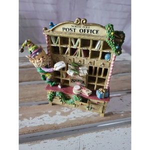 Op de afbeelding: Een miniatuur postkantoor van de Noordpool met een feestelijk elfenbeeld dat post bezorgt. Het postkantoor heeft een houten frame met een rood-wit gestreepte snoepstok, een groene krans en een bord met de tekst "North Pole Post Office". De elf draagt een groene en paarse outfit en houdt een brief vast die geadresseerd is aan "Santa".