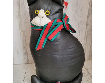 May include: Black plush cat with a white face and yellow eyes wearing a red and green striped scarf. The cat is sitting on a white wooden surface.