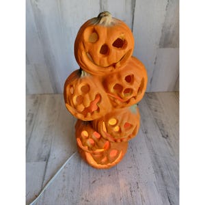 Può includere: Una pila di cinque lanterne di zucca in ceramica arancione con facce intagliate. Le zucche sono illuminate dall'interno, creando un bagliore caldo. Un cavo elettrico bianco si estende dalla base. Una decorazione di Halloween.