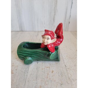 May include: A ceramic planter shaped like a green log with a textured bark finish, featuring a small red elf figure with a pointed hat and upturned feet resting on the top edge of the log. The elf has a painted face with dark eyes and eyebrows.