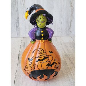 May include: A green witch figurine with purple and black clothing, a black hat with a yellow star, and a swirling orange pumpkin body. The pumpkin has black bats and swirls.