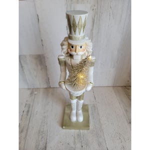 Op de afbeelding: Decoratieve wit en goudkleurige notenkrakerfiguur. De figuur draagt een hoge hoed en houdt een goudkleurige boom met lichtjes vast. De notenkraker staat op een gouden voet.