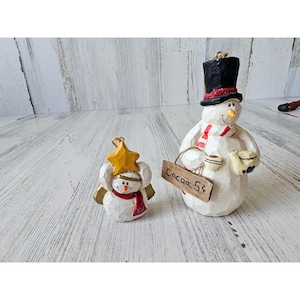 May include: Two white snowman ornaments with painted details. One snowman is wearing a black top hat and a red and white striped scarf. The other snowman is wearing a yellow star on its head and a red scarf. The snowman with the top hat is holding a sign that says "Cocoa 5¢".