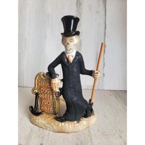 May include: A ceramic figurine of a skeleton wearing a black top hat and a long black coat, leaning against a tombstone that reads GRAVES DUG WHILE YOU WAIT. The skeleton holds a long orange-handled shovel, and the base is textured in tan.
