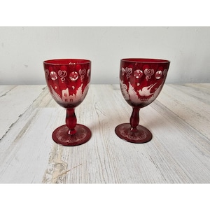 May include: Two ruby red wine glasses with etched white designs. The glasses feature a detailed landscape scene with houses and swirling patterns. The stem and base are also red with etched details. The glasses are on a light wood surface.