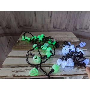 May include: Two strands of Halloween skull string lights. One strand glows green, the other glows white. The skull lights are strung on black wires. The lights are on a weathered, white-painted wooden surface.