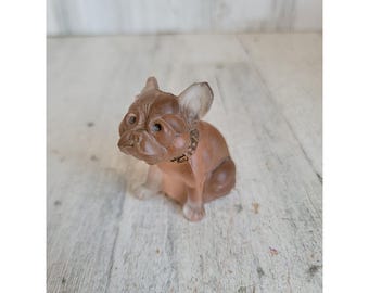 Può includere: Statuetta marrone di un bulldog francese con un piccolo collare. Il cane è seduto con le zampe anteriori distese e la testa leggermente inclinata. La statuetta ha un aspetto liscio e traslucido.