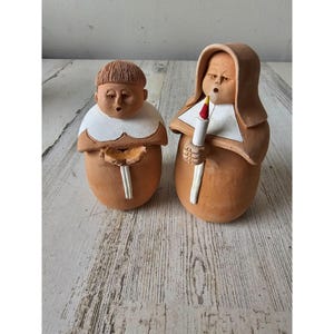 Puede incluir: Dos figuritas de terracota de personas con túnicas, una sosteniendo una vela. Las figuritas están pintadas a mano con detalles blancos y tienen un aspecto rústico y hecho a mano.
