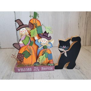 May include: A wooden fall-themed decoration with three pumpkins, two scarecrows, and a black cat. The pumpkins are painted orange with green leaves and plaid accents. The scarecrows are painted in bright colors with hats and straw. The black cat is painted black with white eyes and a pink bow. The base of the decoration has the text "Welcome to our Patch".