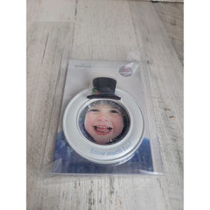 以下が含まれることがあります： 黒いシルクハットをかぶった白い雪だるまの形をした写真フレーム。中央には子供の顔が写っています。フレームには「Snow much fun!」と書かれています。