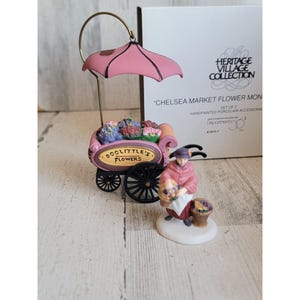 Könnte beinhalten: Eine Miniatur-Blumenkarre aus Porzellan mit rosa Baldachin und dem Text "DOOLITTLE'S FLOWERS". Eine Frau in einem rosa Mantel und Hut hält einen Blumenkorb. Teil der "Heritage Village Collection".