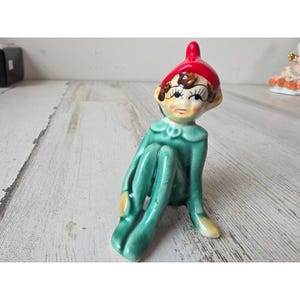 May include: A ceramic figurine of a seated elf with a red cap and green outfit. The elf has a whimsical expression with painted facial features. The figurine is on a light-colored wooden surface.