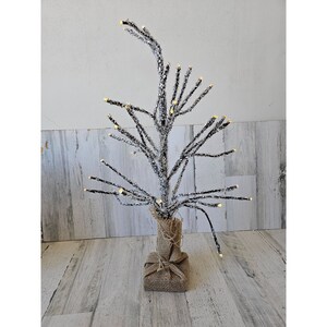 May include: A snowy, twig-like artificial Christmas tree with warm white LED lights. The tree is in a burlap wrapped base.