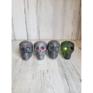 May include: Four decorative skull figurines with different colored eyes. The skulls are made of a dark gray material and have a textured surface. The eyes are red, green, and black.
