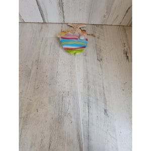 May include: A decorative garden stake featuring a flat, painted metal bunny head emerging from a colorful, striped egg shape. The egg displays horizontal bands of blue, pink, and green, attached to a long, thin, white metal rod for insertion.