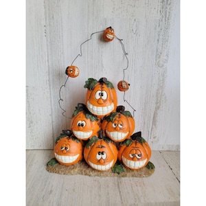 May include: A whimsical Halloween decoration featuring a stack of orange pumpkins with smiling faces. A wire arch holds smaller pumpkins. The pumpkins have green leaves and are set on a brown base. A fun, festive accent for the fall season.