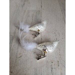 Puede incluir: Dos adornos decorativos de pájaros blancos con detalles de purpurina plateada y largas colas de plumas blancas y esponjosas. Cada pájaro tiene un clip plateado para sujetarlo. Sobre una superficie de madera clara.