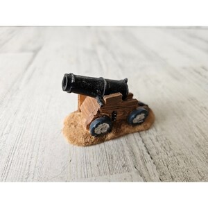 May include: A miniature black cannon on a wooden base with wheels. The cannon is mounted on a small pile of sand.