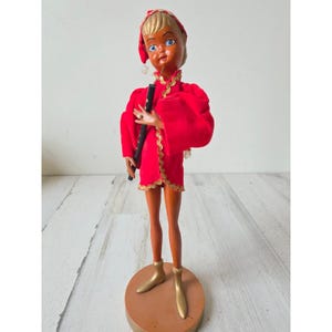May include: A vintage doll with a painted face, wearing a red velvet outfit with gold trim, a matching cap, and gold boots. The doll is holding a black flute and stands on a brown circular base. The doll's skin is tan and it has blue eyes.