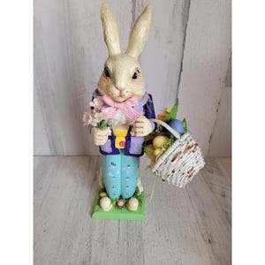 Puede incluir: Figura decorativa de conejito de Pascua. El conejito lleva una chaqueta morada, pantalones azules y una pajarita rosa. Sostiene un ramo de flores rosas y una cesta llena de huevos y flores de Pascua. La figura está sobre una base verde.