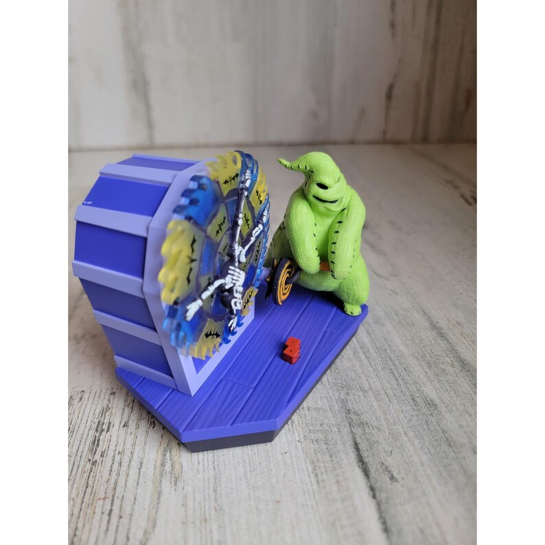 May include: A green, stitched, Nightmare Before Christmas character, known as the "Boogeyman", is holding a spinning wheel with a skeleton design. The wheel is attached to a purple and blue base.