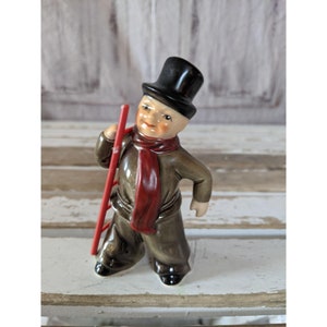May include: A ceramic figurine of a chimney sweep wearing a black top hat and a red scarf, holding a red ladder.
